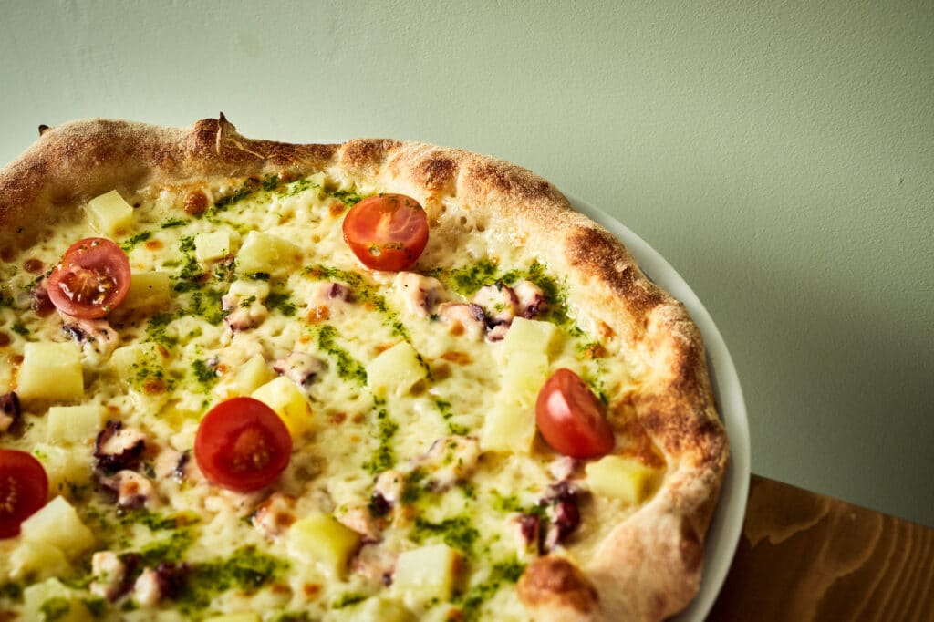 Prawdziwa włoska pizza bianca z ośmirniczkami, kawałkami długodojrzewającego sera oraz pomidorkami koktailowymi na oliwkowym tle.