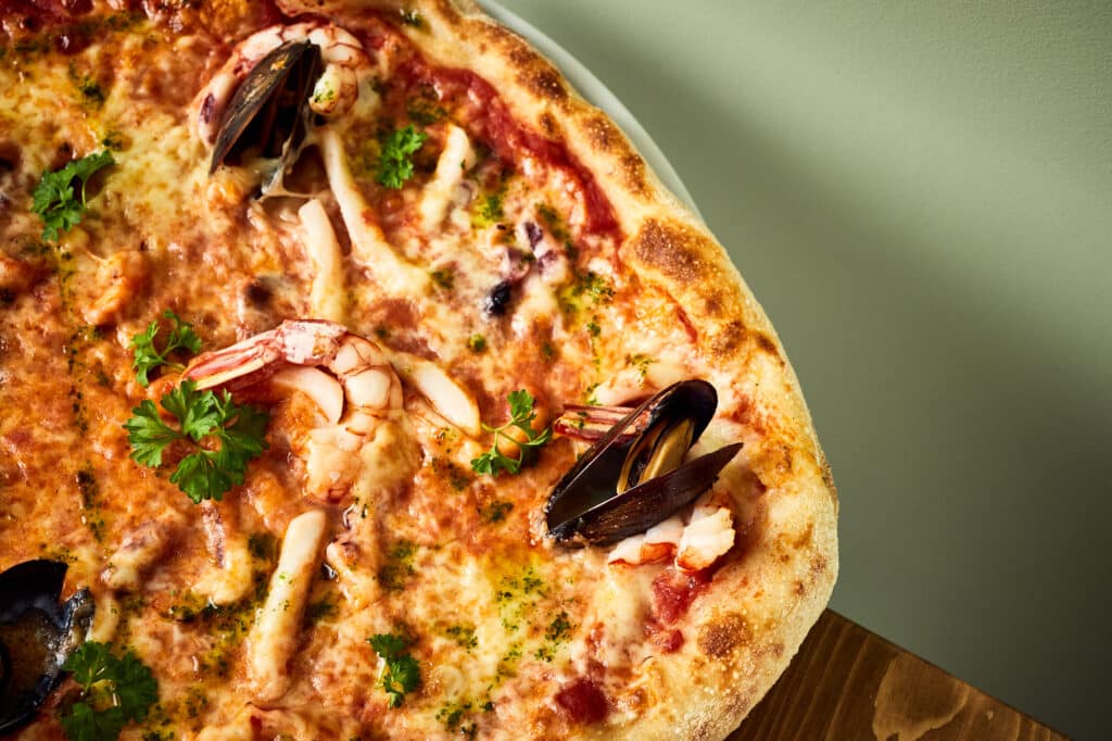 Zbliżenie na pizzę z owocami morza. Widać owoce morza zanurzone w kremowym serze wyłożonym na sosie pomidorowym.