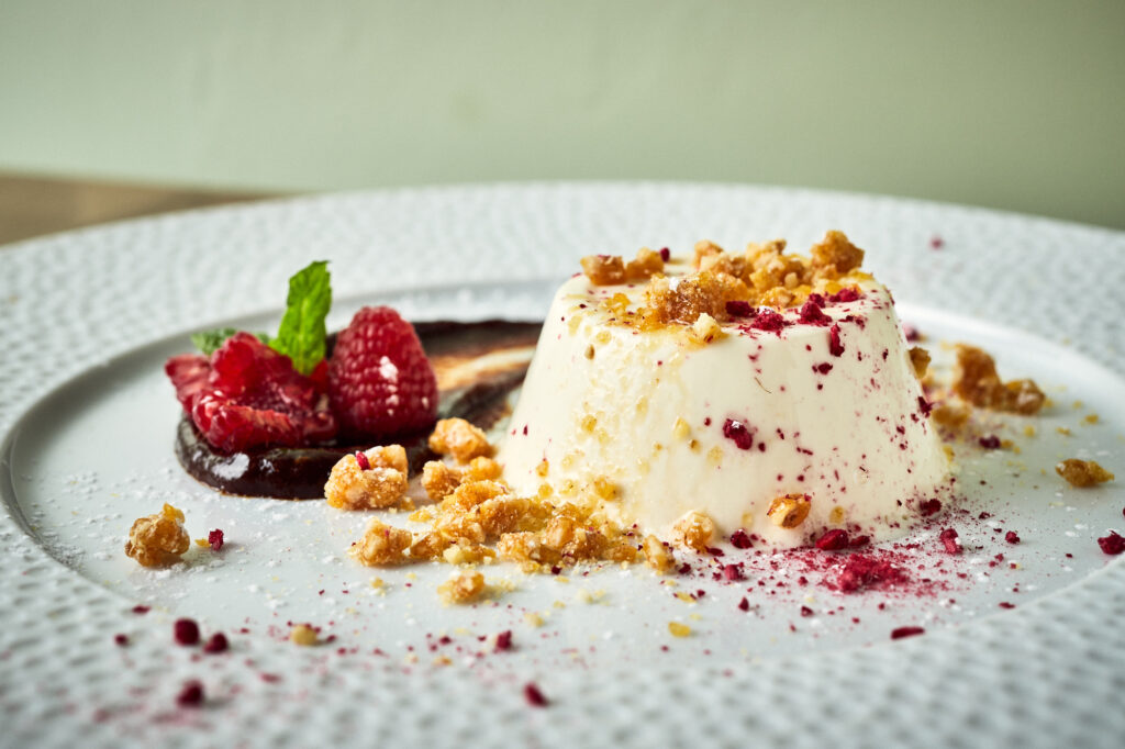 Włoska panna cotta podana na białym, porcelanowym talerzu z prażonymi orzechami i świeżymi malinami.