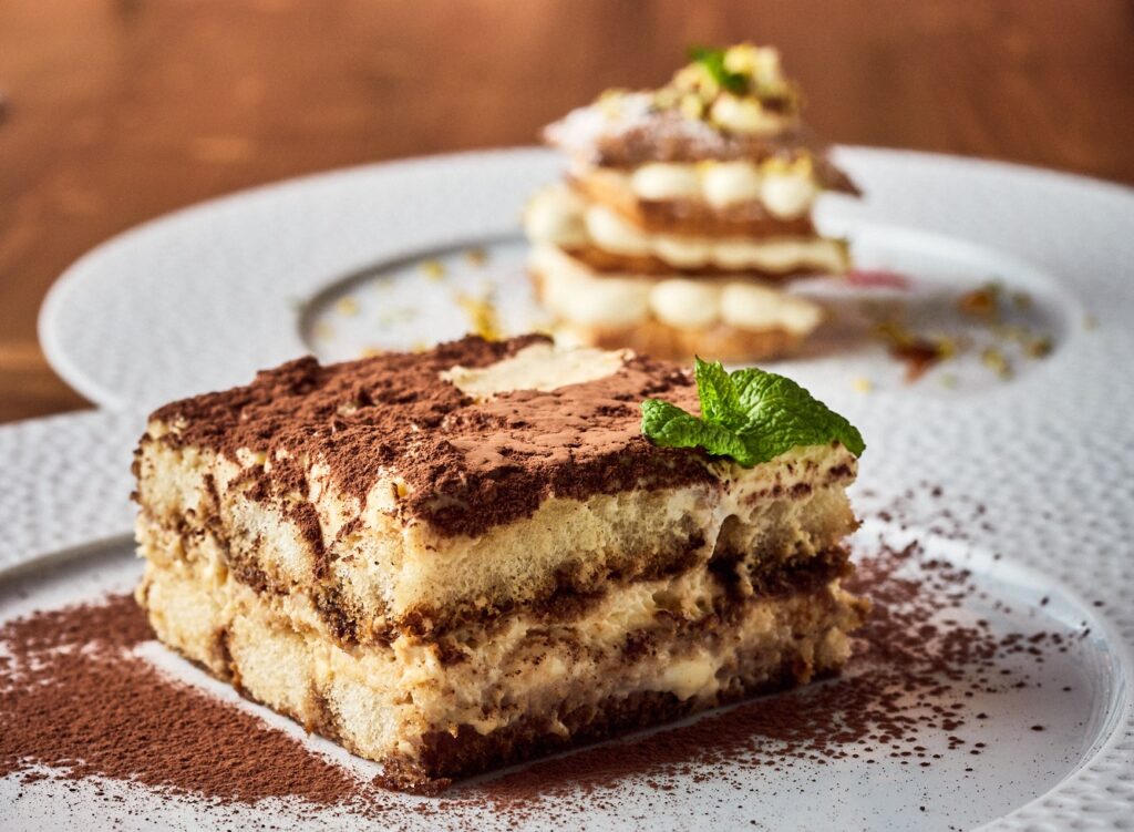 Włoski deser - klasyczne tiramisu podane na białym porcelanowym talerzu  obsypanym kakao. Ciasto jest podane z listkami mięty.