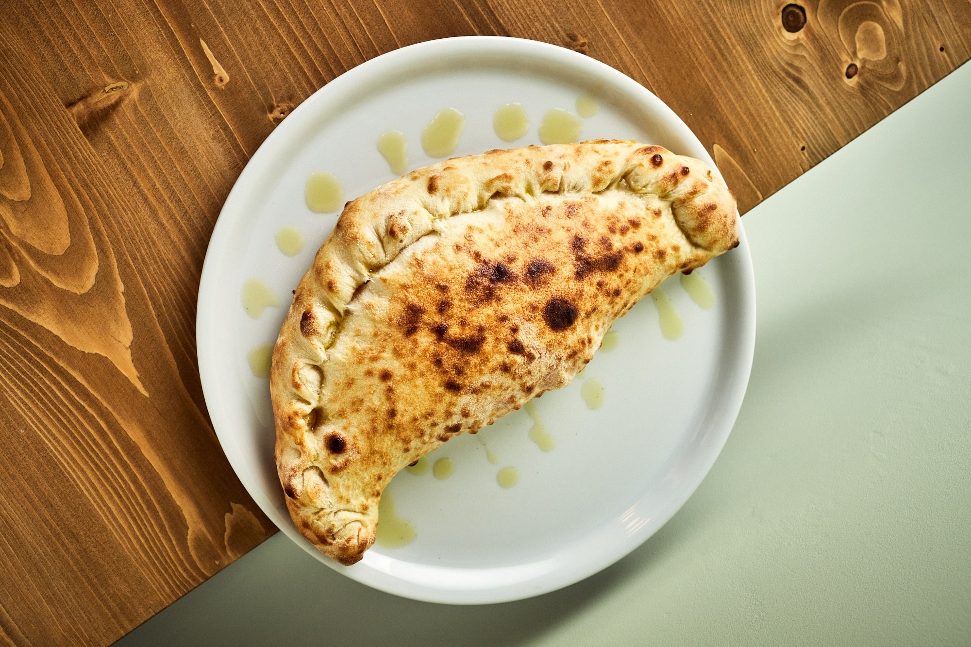Calzone na białym, porcelanowym talerzu, położonym na drewnianym stole. Talerz dokoła pieroga jest pokropiony oliwą.