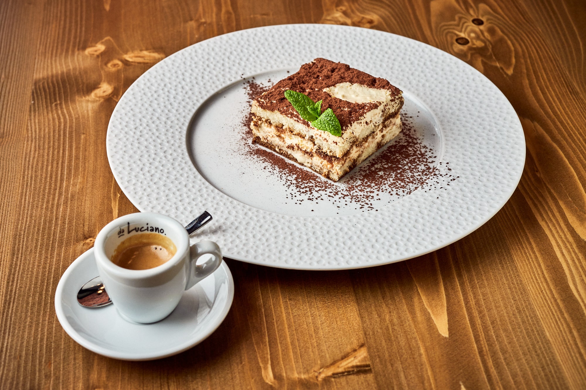 Włoska kawa espresso w filiżance oraz klasyczne tiramisu podane na ozdobnym białym talerzu, ustawione na drewnianym stole.